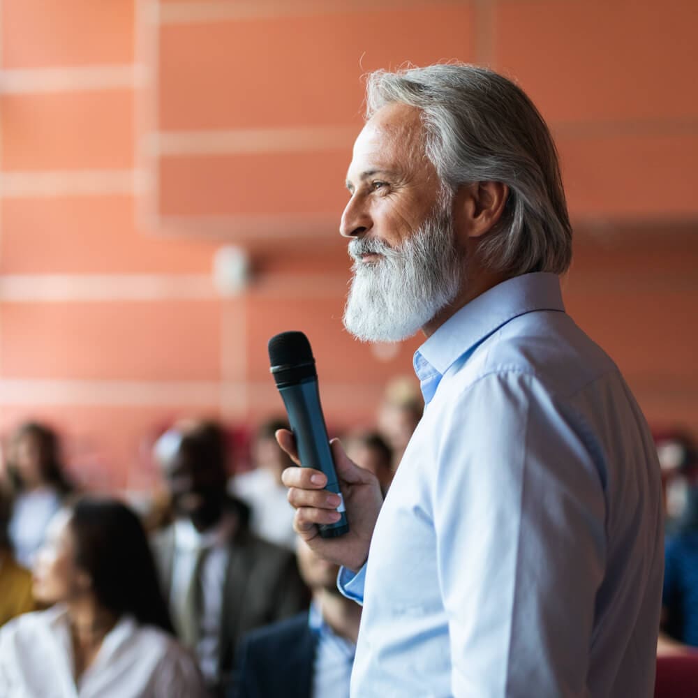 A bearded man holds a microphone and is answering questions in front of a crowd during a presentation.