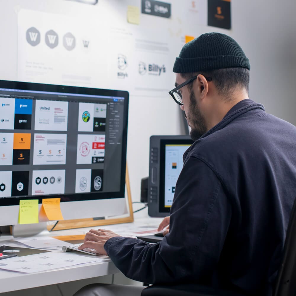 A man in a beanie sits at a computer and edits slides.