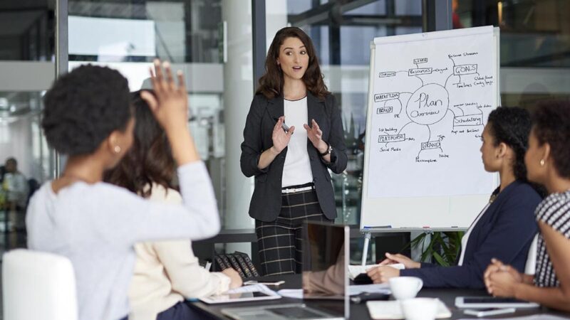 A woman leads a presentation in front of a conference room of people.