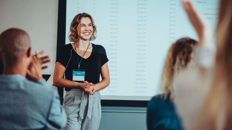 A person at the front of a conference room leading a presentation is smiling, while attendees clap.